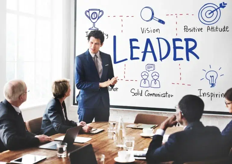 A person in a suit presents to a group in a conference room. The whiteboard displays the word "LEADER" surrounded by diagrams with traits like "Vision" and "Inspiration."