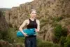 Woman in athletic wear holding a blue yoga mat stands in front of rocky cliffs, conveying a sense of calm and focus amidst nature.