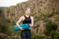 Woman in athletic wear holding a blue yoga mat stands in front of rocky cliffs, conveying a sense of calm and focus amidst nature.