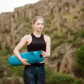 Woman in athletic wear holding a blue yoga mat stands in front of rocky cliffs, conveying a sense of calm and focus amidst nature.