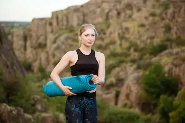 Woman in athletic wear holding a blue yoga mat stands in front of rocky cliffs, conveying a sense of calm and focus amidst nature.