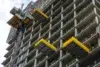 A high-rise concrete building under construction with yellow platforms extended from its sides and a construction lift on the left.