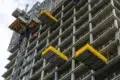 A high-rise concrete building under construction with yellow platforms extended from its sides and a construction lift on the left.