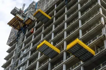 A high-rise concrete building under construction with yellow platforms extended from its sides and a construction lift on the left.