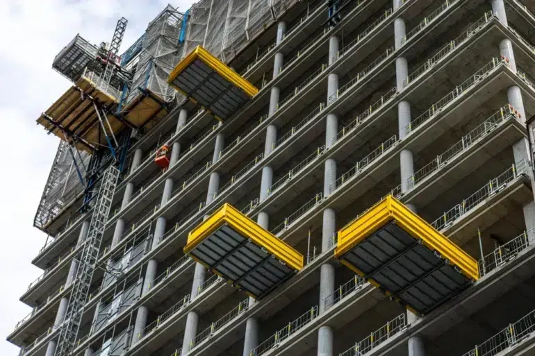 A high-rise concrete building under construction with yellow platforms extended from its sides and a construction lift on the left.