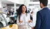 A woman stands next to a yellow car, smiling and talking to a man in a suit who is holding a clipboard inside a car dealership.