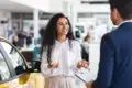 A woman stands next to a yellow car, smiling and talking to a man in a suit who is holding a clipboard inside a car dealership.
