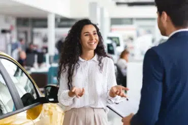 A woman stands next to a yellow car, smiling and talking to a man in a suit who is holding a clipboard inside a car dealership.