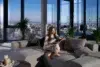 A woman sits on a gray sectional sofa, reading in a modern apartment with large windows offering a city view. The room is bright and serene, with plants nearby.