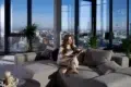 A woman sits on a gray sectional sofa, reading in a modern apartment with large windows offering a city view. The room is bright and serene, with plants nearby.