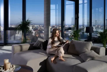 A woman sits on a gray sectional sofa, reading in a modern apartment with large windows offering a city view. The room is bright and serene, with plants nearby.