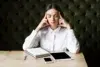 Woman in a white shirt sitting at a wooden table with her eyes closed. She touches her temples, appearing thoughtful. Nearby are a notebook, tablet, and phone.