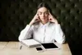 Woman in a white shirt sitting at a wooden table with her eyes closed. She touches her temples, appearing thoughtful. Nearby are a notebook, tablet, and phone.