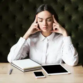 Woman in a white shirt sitting at a wooden table with her eyes closed. She touches her temples, appearing thoughtful. Nearby are a notebook, tablet, and phone.