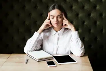 Woman in a white shirt sitting at a wooden table with her eyes closed. She touches her temples, appearing thoughtful. Nearby are a notebook, tablet, and phone.