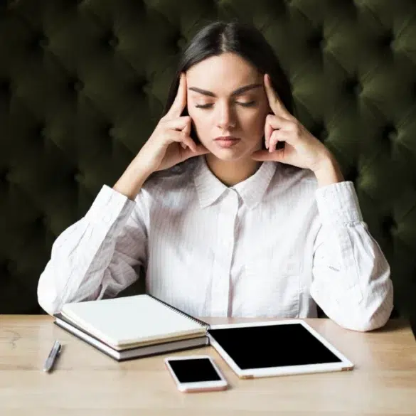 Woman in a white shirt sitting at a wooden table with her eyes closed. She touches her temples, appearing thoughtful. Nearby are a notebook, tablet, and phone.