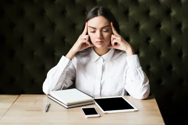 Woman in a white shirt sitting at a wooden table with her eyes closed. She touches her temples, appearing thoughtful. Nearby are a notebook, tablet, and phone.