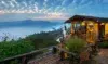 Cozy wooden cabin on a hillside with people enjoying a misty mountain sunrise and lush greenery surrounding the area.