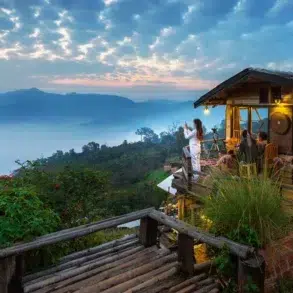 Cozy wooden cabin on a hillside with people enjoying a misty mountain sunrise and lush greenery surrounding the area.