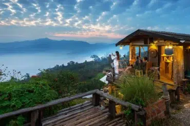 Cozy wooden cabin on a hillside with people enjoying a misty mountain sunrise and lush greenery surrounding the area.