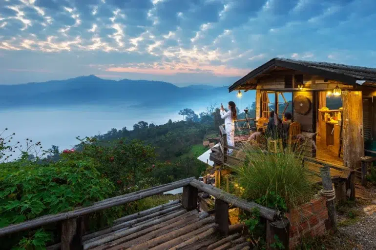 Cozy wooden cabin on a hillside with people enjoying a misty mountain sunrise and lush greenery surrounding the area.