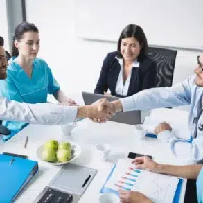 A doctor and a businessman shake hands across a table in a meeting. Colleagues in scrubs and formal attire watch, with charts and apples on the table.