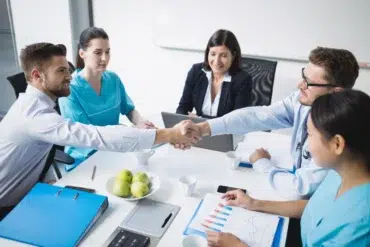 A doctor and a businessman shake hands across a table in a meeting. Colleagues in scrubs and formal attire watch, with charts and apples on the table.