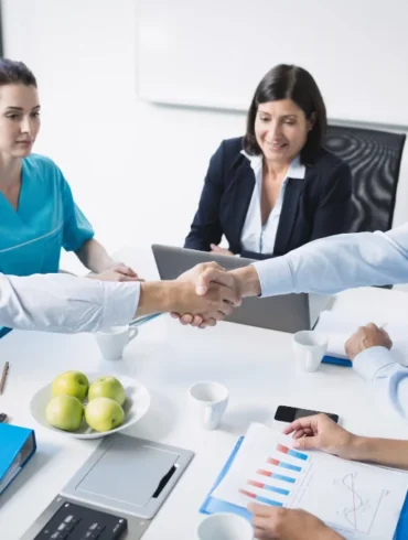 A doctor and a businessman shake hands across a table in a meeting. Colleagues in scrubs and formal attire watch, with charts and apples on the table.