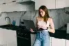 Woman in jeans and white tank top using a laptop in a modern kitchen.