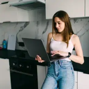 Woman in jeans and white tank top using a laptop in a modern kitchen.