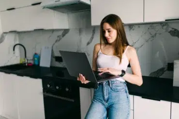 Woman in jeans and white tank top using a laptop in a modern kitchen.