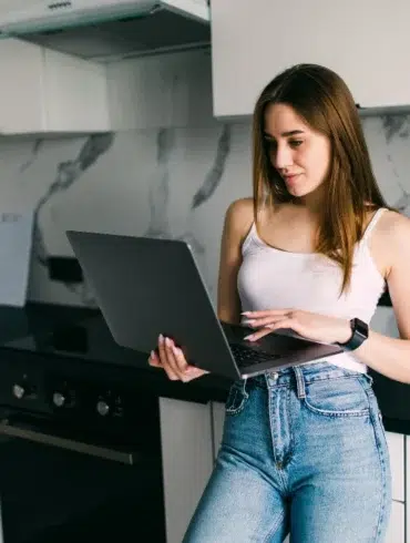Woman in jeans and white tank top using a laptop in a modern kitchen.