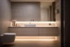 A modern bathroom featuring a sleek vanity with undermount lighting, a white sink, and a minimalistic design; a toilet is positioned alongside the vanity against gray walls.