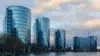 Five modern, curved glass office buildings reflecting the cloudy sky, situated along a waterfront with leafless trees nearby.
