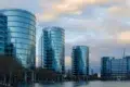 Five modern, curved glass office buildings reflecting the cloudy sky, situated along a waterfront with leafless trees nearby.