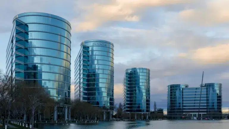 Five modern, curved glass office buildings reflecting the cloudy sky, situated along a waterfront with leafless trees nearby.