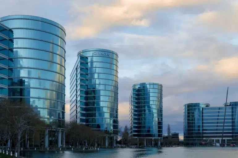 Five modern, curved glass office buildings reflecting the cloudy sky, situated along a waterfront with leafless trees nearby.