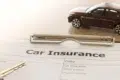 A car insurance form lies on a table with a pen and a car key nearby. A small toy car rests on the form, symbolizing automobile coverage.