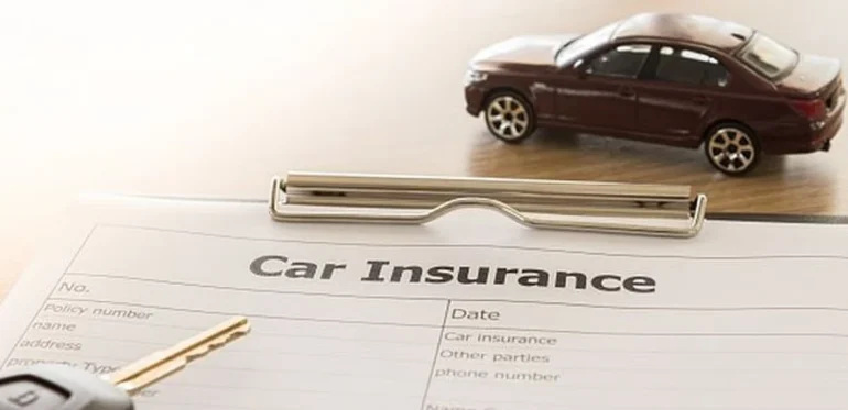A car insurance form lies on a table with a pen and a car key nearby. A small toy car rests on the form, symbolizing automobile coverage.