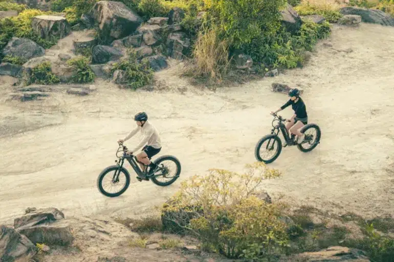 Two cyclists wearing helmets ride electric mountain bikes on a rocky, dirt trail surrounded by greenery, conveying adventure and outdoor exploration.