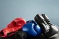 Three pairs of boxing gloves—red, blue, and black—are stacked on a dark surface against a plain light blue background, symbolizing sports and competition.