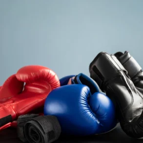 Three pairs of boxing gloves—red, blue, and black—are stacked on a dark surface against a plain light blue background, symbolizing sports and competition.