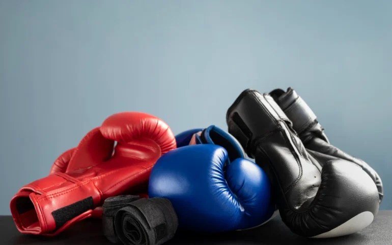 Three pairs of boxing gloves—red, blue, and black—are stacked on a dark surface against a plain light blue background, symbolizing sports and competition.
