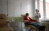 A nurse in a mask and gloves stands near a patient in a recliner receiving an IV drip in a brightly lit medical room.