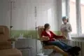 A nurse in a mask and gloves stands near a patient in a recliner receiving an IV drip in a brightly lit medical room.