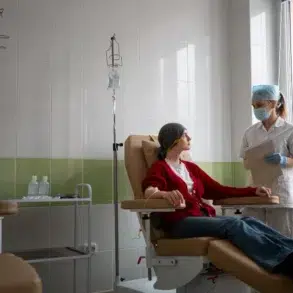 A nurse in a mask and gloves stands near a patient in a recliner receiving an IV drip in a brightly lit medical room.