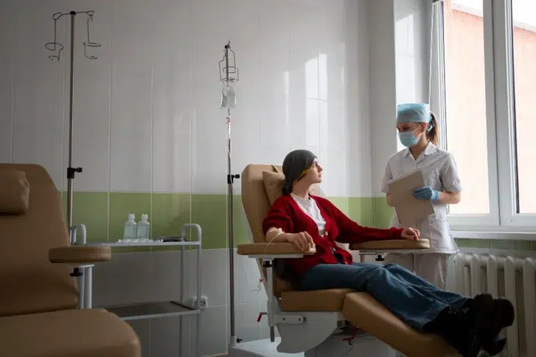 A nurse in a mask and gloves stands near a patient in a recliner receiving an IV drip in a brightly lit medical room.
