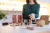 A woman in a green blouse organizes assorted beauty products, including tubes, bottles, and brushes on a white table. The setting appears modern and well-lit.