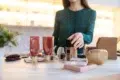 A woman in a green blouse organizes assorted beauty products, including tubes, bottles, and brushes on a white table. The setting appears modern and well-lit.