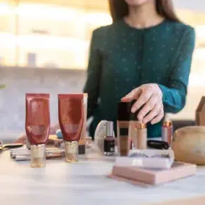 A woman in a green blouse organizes assorted beauty products, including tubes, bottles, and brushes on a white table. The setting appears modern and well-lit.